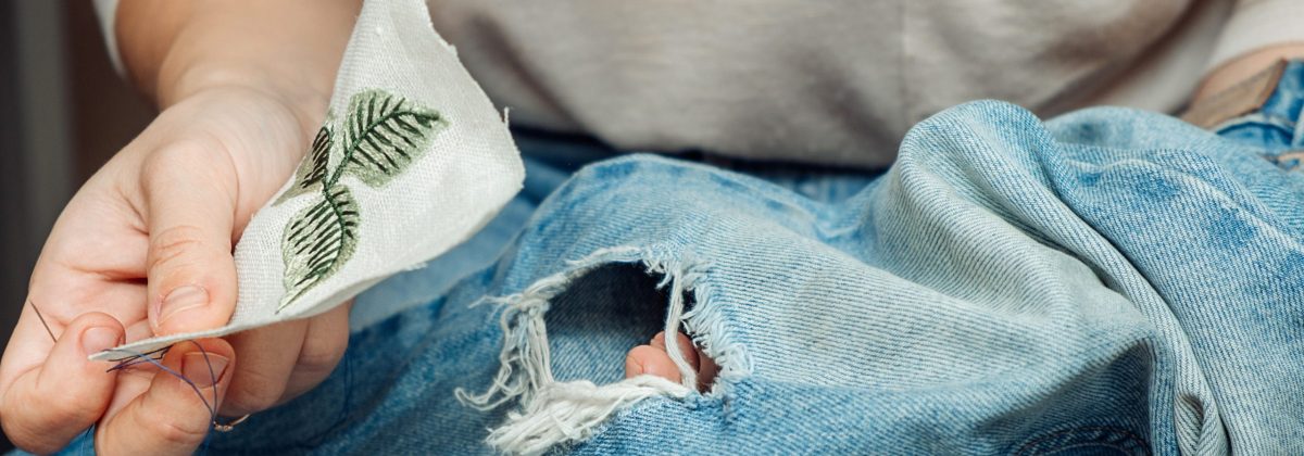 A woman mends jeans, sews a patch on a hole, hands close-up.Mending clothes concept,reusing old jeans